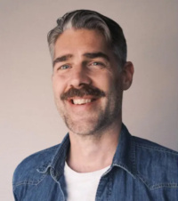 Ein Porträtfoto des Interviewteilnehmers mit blauem Hemd, weißem T-Shirt und grauen Haaren.