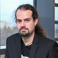 Porträtfoto eines MAnnes mit langen Haaren, Bart und einem schwarzen Jackett.