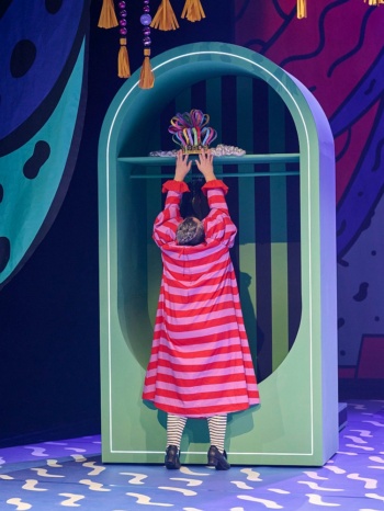 Eine Person in einem rosafarben geringelten Kleid steht mit dem Rücken zur Kamera und streckt sich nach einem großen bunten Hut, der im oberen Fach eines Schrankes liegt.