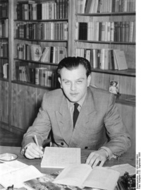 Ein Mann im zweireihigen Anzug sitzt an einem Tisch und hält Block und Stift in der Hand. Hinter ihm ist eine Bücherwand.