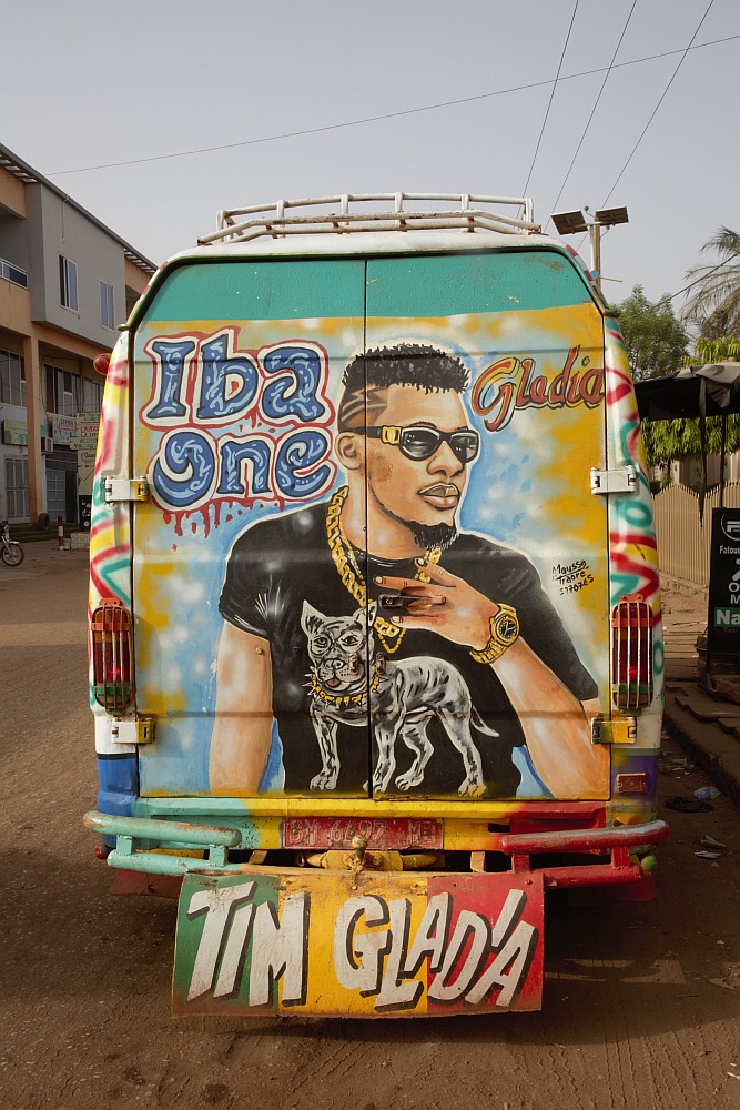 Ein Kleinbus auf einer Straße in einer Afrikanischen Ortschaft. Auf der Rückseite ist das übergroße Porträt eines Sängers zu sehen.
