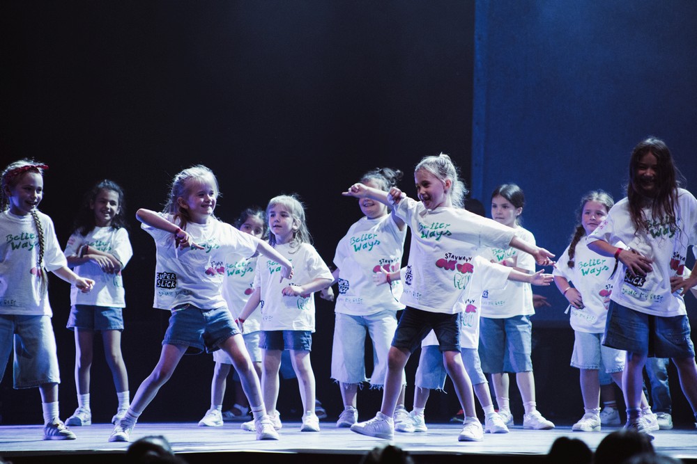 Kinder in weißen Shirts und Jeans bei einer Tanzchoreographie im Rampenlicht auf einer ansonsten dunklen Bühne.