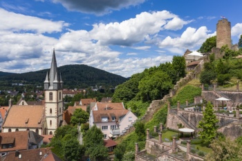 Eine kleine Stadt in einem Bergtal. Im Zentrum liegt eine Kirche, am Berghang eine Burgruine.