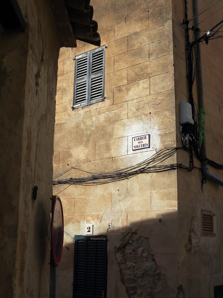Eine Gasse in einer kleinen mediterranene Ortschaft. an den Hauswänden verlaufen Stromkabel, darüber hängt ein Straßenschild.