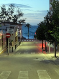 Eine leicht abschüssige gerade Straße in einer Ortschaft, die direkt auf das Meer hinführt. Am Ende der Straße steht eine Palme am Wasser.