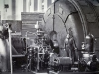 Eine historische Aufnahme aus einem Wasserkraftwerk. Menschen stehen neben großen Turbinen.