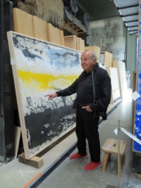 Ein Maler in schwarzer Kleidung steht in einem Atelier und zeigt auf ein großes Gemälde