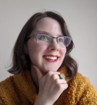 Porträtfoto einer Frau mit brauenen Haaren, roten Lippen, einem gelben Pullover und einem großen grünen Ring am Finger.