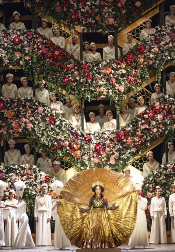 Zahlreiche Schauspieler auf einer mit vielen Blumen geschmückten Bühne. Die Hauptdarstellerin trägt ein goldenes Kleid