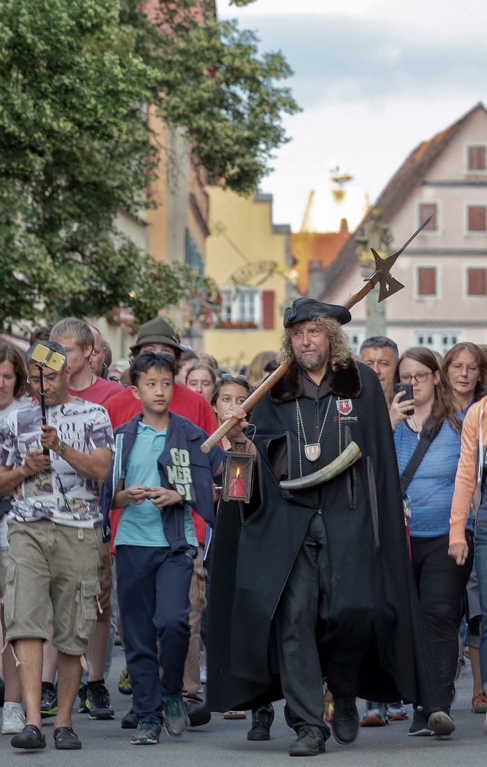 Ein Nachtwächter mit schwarzer Kutte, Laterne und Hellebarde führt eine große Gruppe von Menschen durch eine Straße