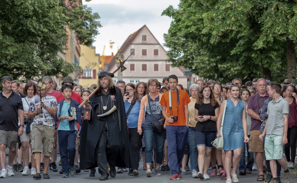 Ein Nachtwächter mit schwarzer Kutte, Laterne und Hellebarde führt eine große Gruppe von Menschen durch eine Straße