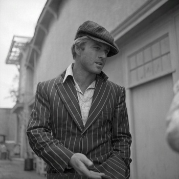 Robert Redford auf einer schwarz-weiß- Aufnahme. Er trägt einen Nadelstreifenanzug mit offenem Hemd und Schiebermütze.