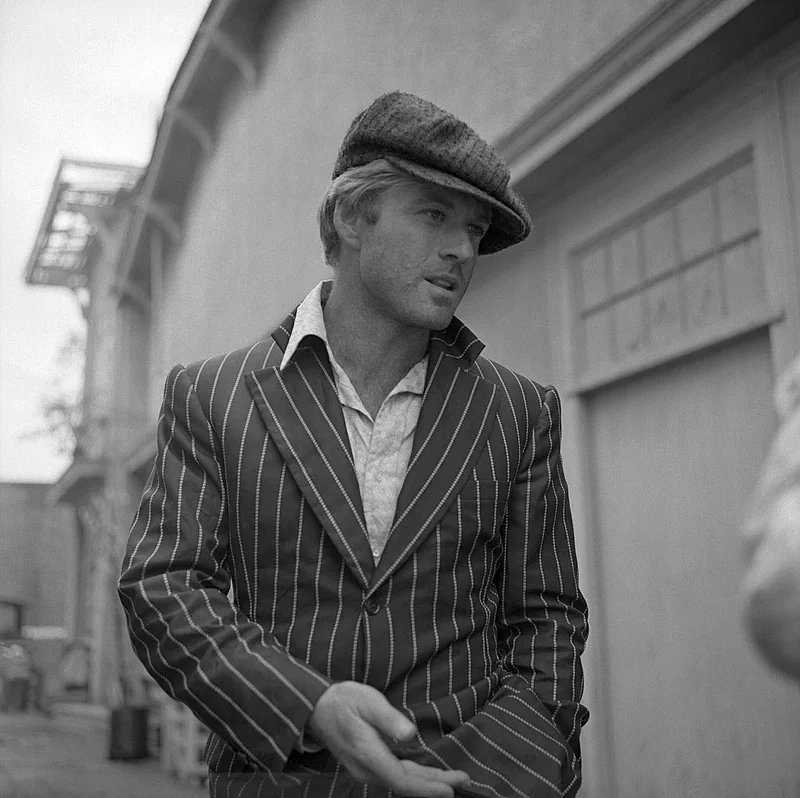 Robert Redford auf einer schwarz-weiß- Aufnahme. Er trägt einen Nadelstreifenanzug mit offenem Hemd und Schiebermütze.