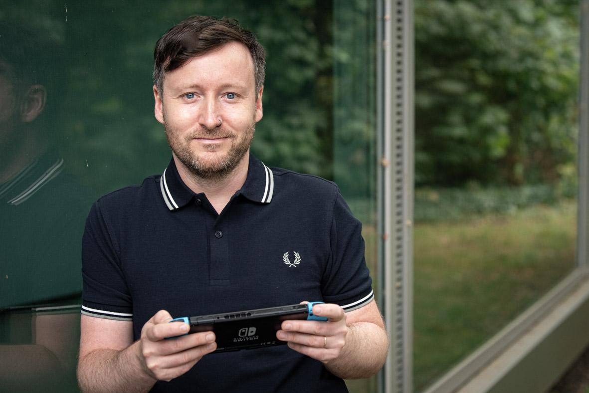 Ein Mann mit braunen Haaren und hellen Augen, der ein Poloshirt trägt und ein Videospielgerät in den Händen hält.