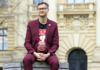 Der Autor sitzt auf einer Holzbank und trägt einen dunkelroten Anzug. Seine Hände liegen verschränkt auf seinem Schoß.