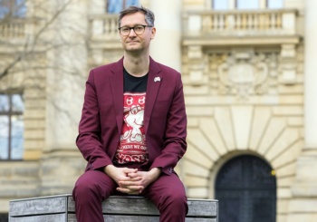 Der Autor sitzt auf einer Holzbank und trägt einen dunkelroten Anzug. Seine Hände liegen verschränkt auf seinem Schoß.