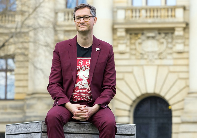 Der Autor sitzt auf einer Holzbank und trägt einen dunkelroten Anzug. Seine Hände liegen verschränkt auf seinem Schoß.
